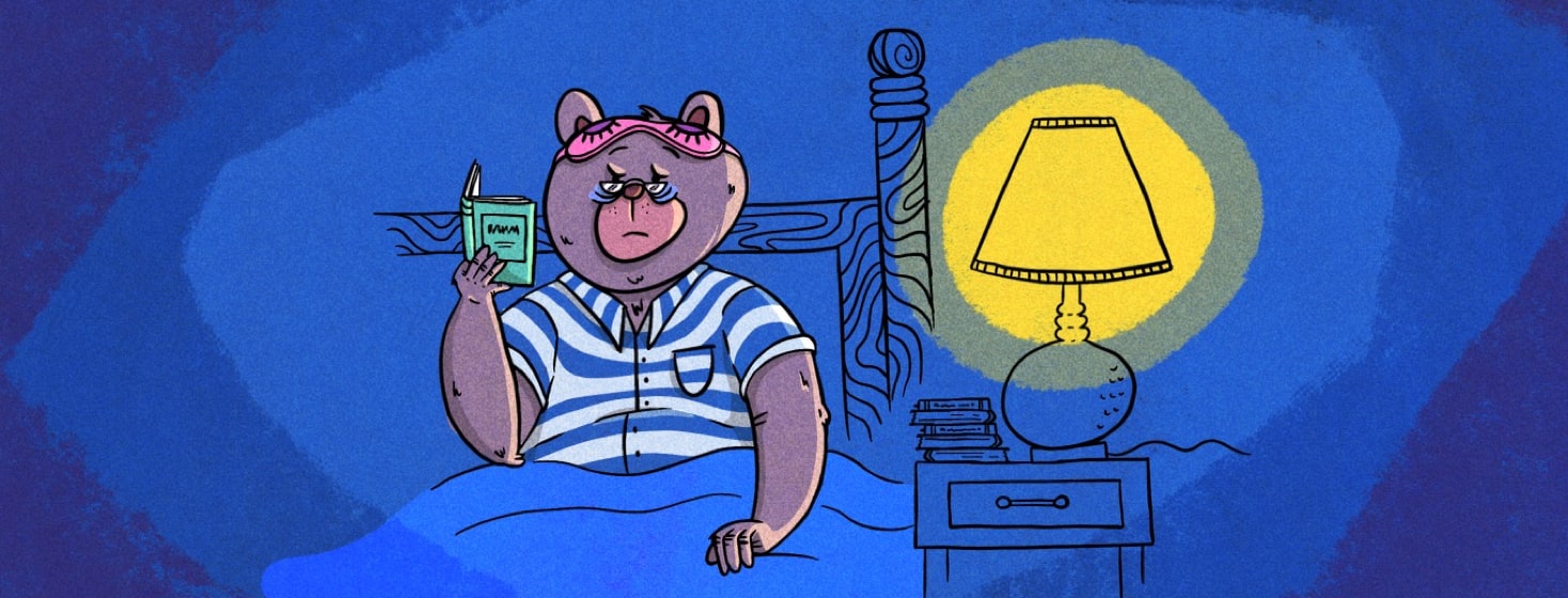 A bear in pajamas sitting in bed with an eye mask propped on top of his head reading a book in the lamplight